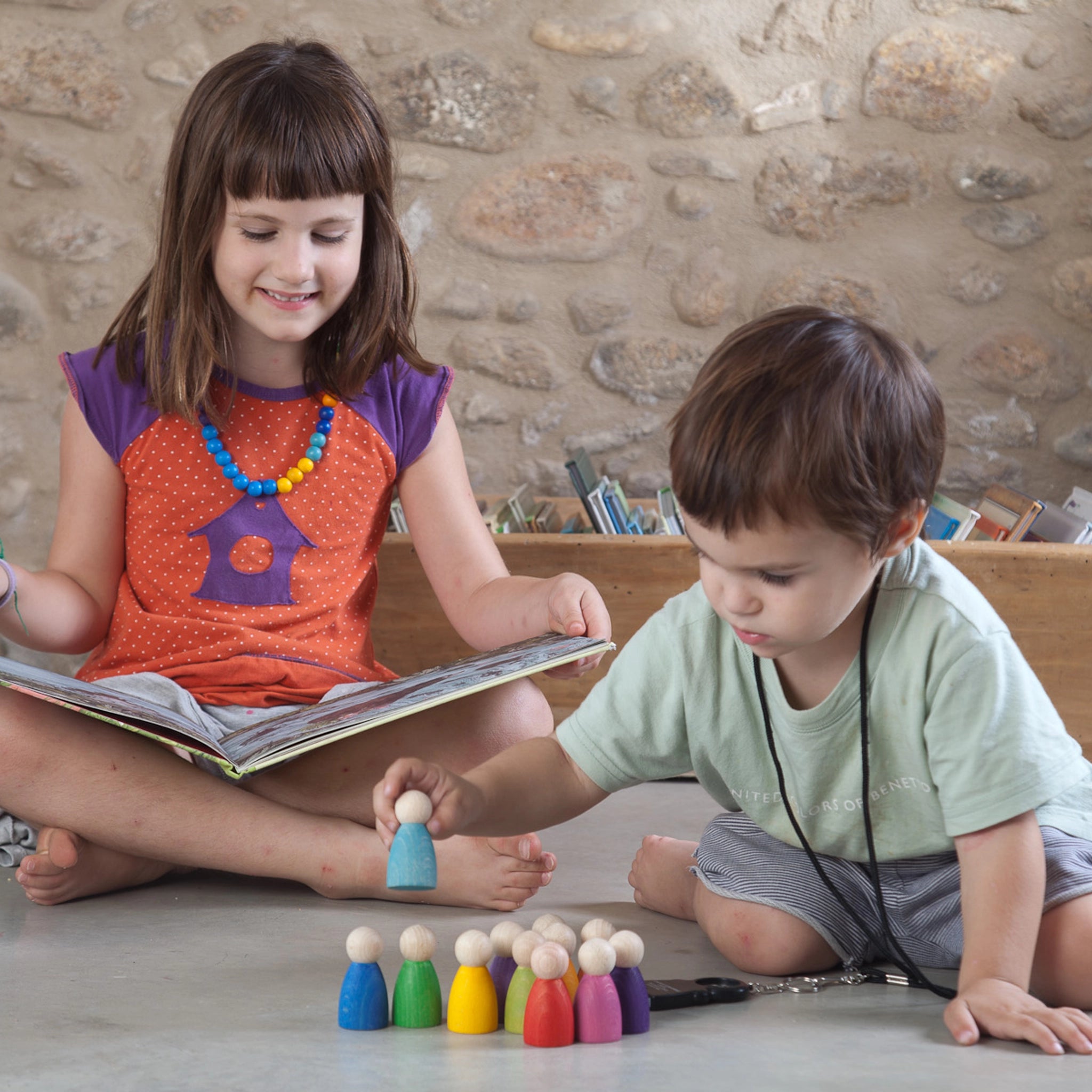 Freies Spiel: 12 Nins aus nachhaltig geforstetem Holz, handgefertigt in Spanien. Zwei Kinder spielen mit einem Buch, umgeben von farbenfrohen Nins-Figuren, ideal für kreatives Spiel.