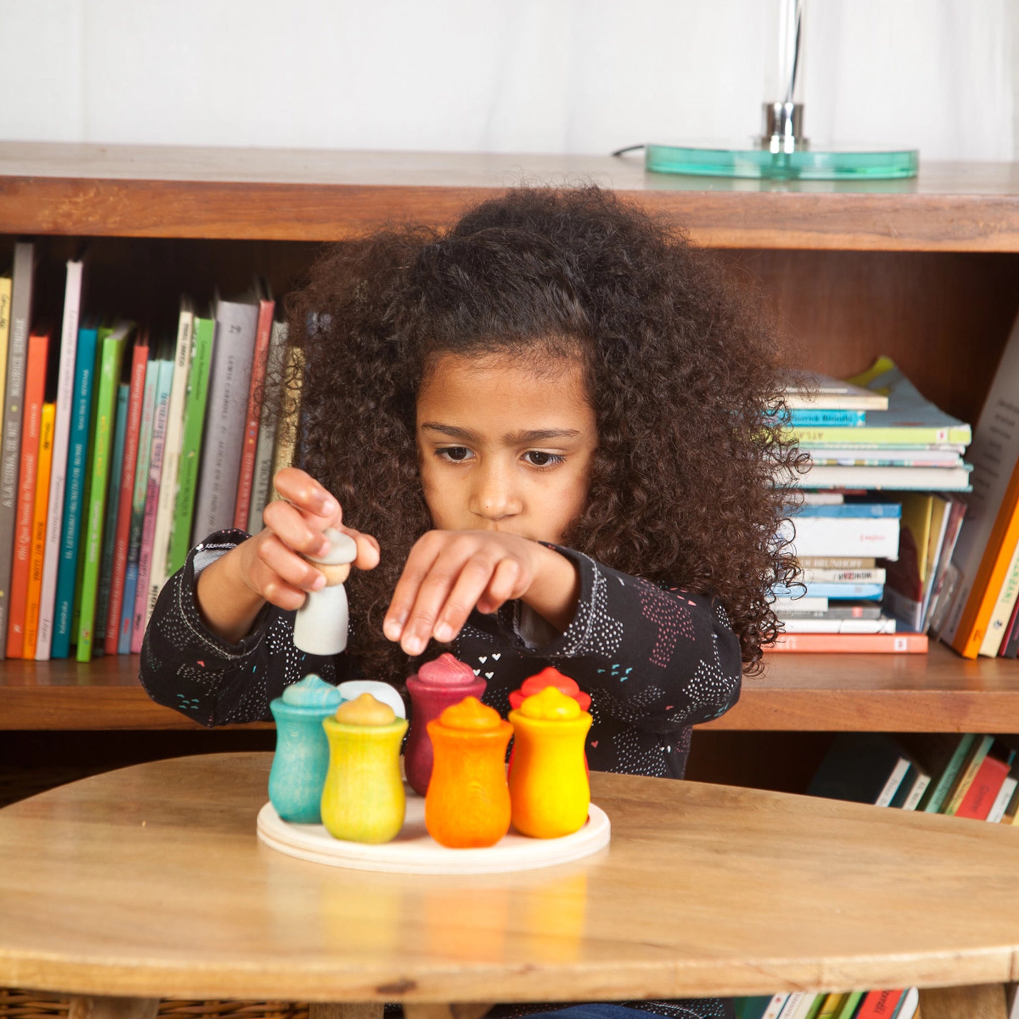 Freies Spiel: Weekly Calendar, ein handgefertigtes Set aus Holzfiguren und Töpfchen, wird von einem Mädchen auf einem Tisch erkundet. Ideal zur Förderung des Wochentagsverständnisses bei Kindern.