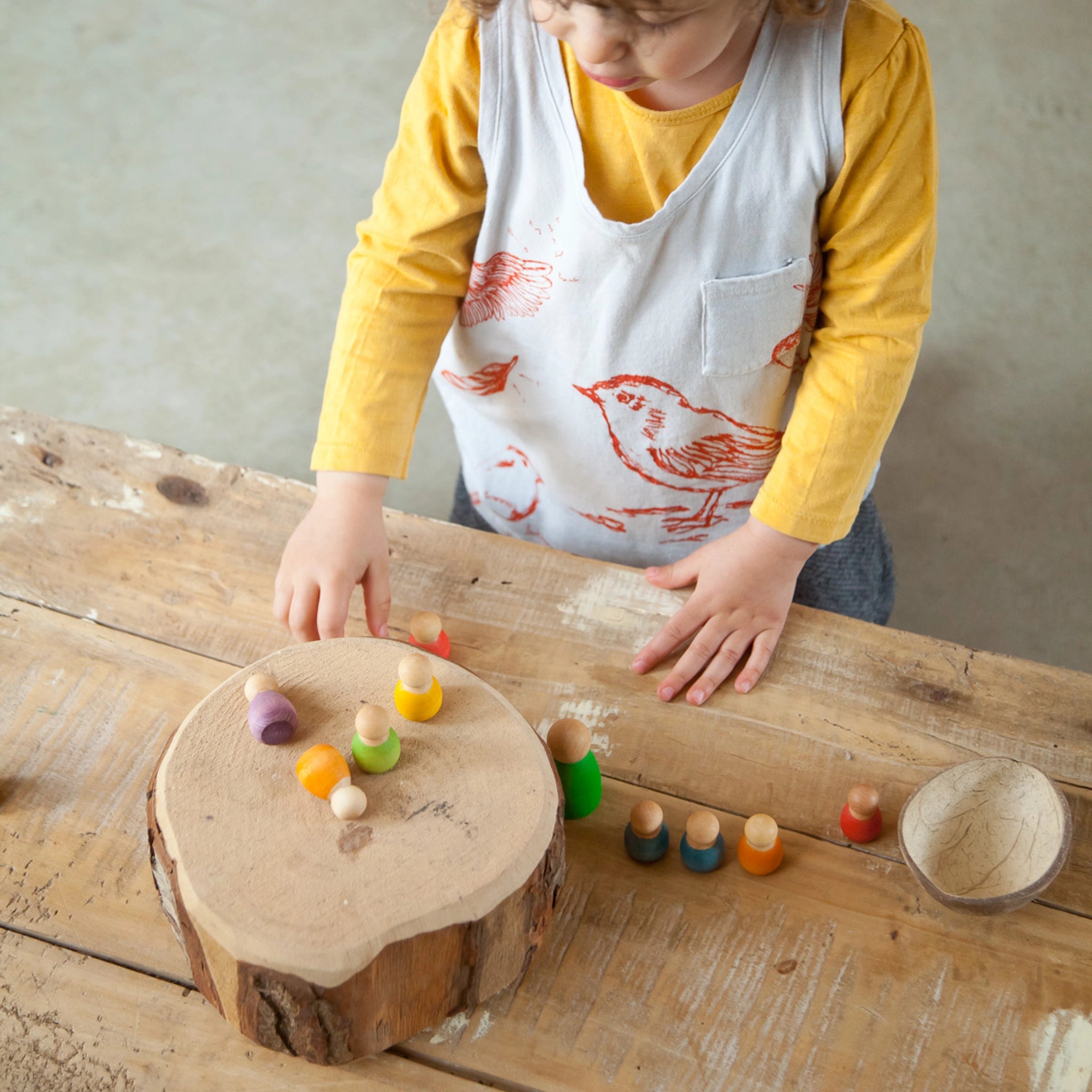 Freies Spiel: Baby Nins, ein Kind spielt mit handgefertigten Holzfiguren auf einem Baumstumpf. Die sechs Figuren aus nachhaltig geforstetem Holz sind in einer Kokosnussschale arrangiert.