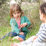 Zwei Kinder sitzen im Gras; eines hält einen Stein. Freies Spiel: Dark Adult Nins, handgefertigt aus dunklem Holz, perfekt für kreatives, nachhaltiges Spiel.