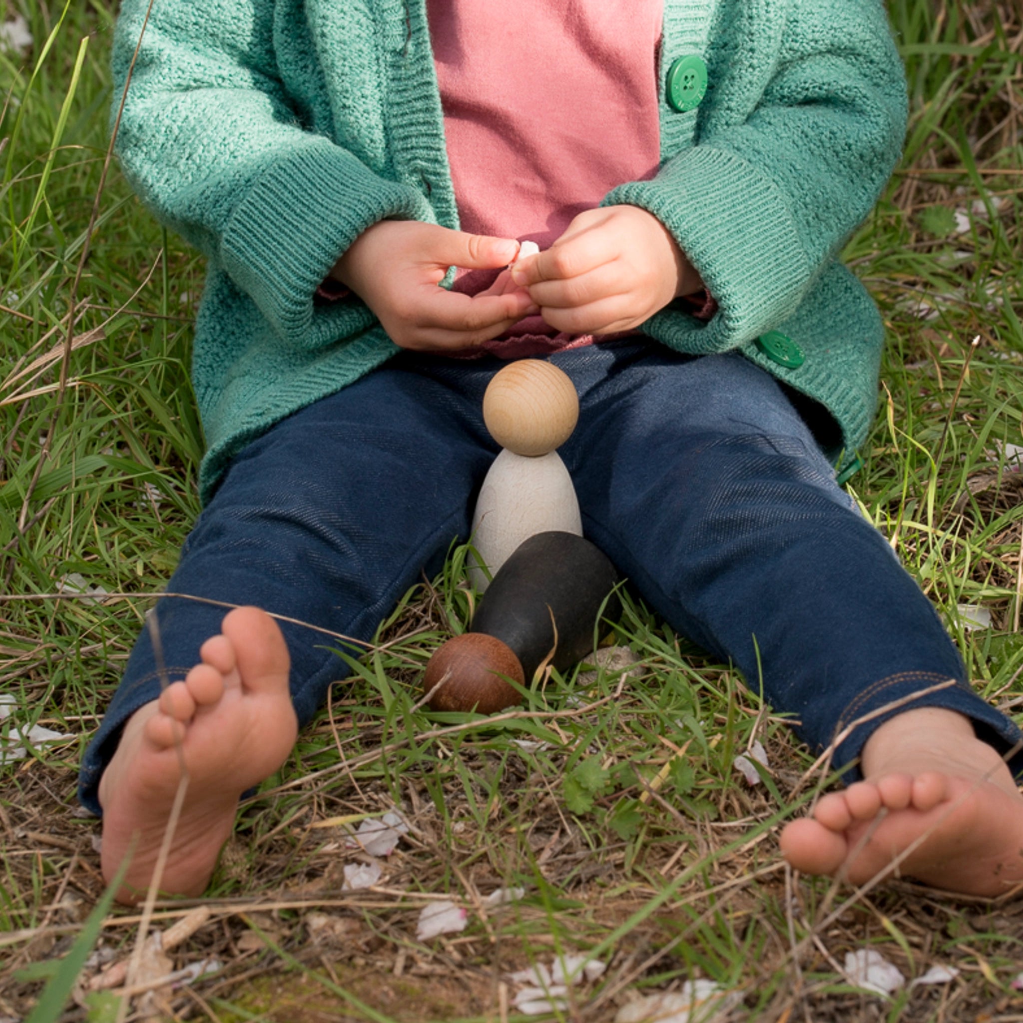 Freies Spiel: Dark Adult Nins, handgefertigte Holzfiguren, sitzen in Gras neben einem Kind. Ideal für freies Spiel, nachhaltig in Spanien produziert, kombinierbar mit Grapat Nins.