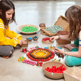 Zwei Mädchen spielen auf dem Boden mit Freies Spiel: Mandala Fire, einem handgefertigten Set aus 36 roten Holz-Feuerelementen, ideal für kreative Mandalas und Landschaften.