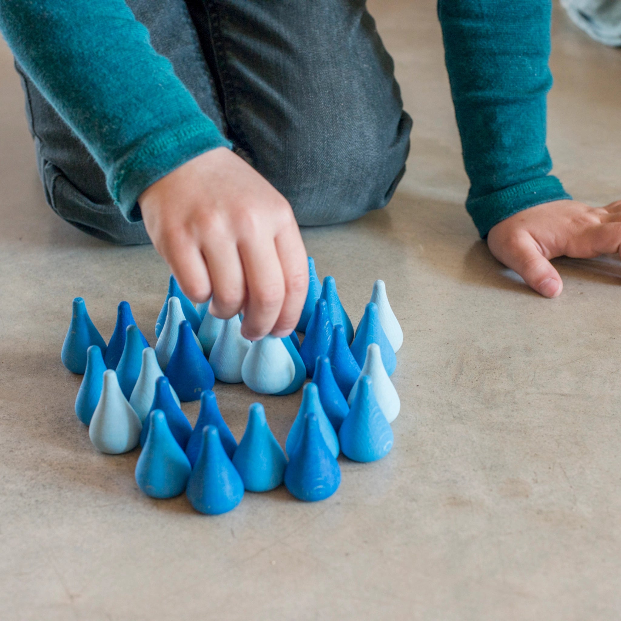 Freies Spiel: Mandala Raindrop – Ein Kind spielt mit handgefertigten blauen Regentropfen aus Holz, ideal für kreative Kunstwerke und Landschaften. Nachhaltig produziert und perfekt für fantasievolles Spiel.