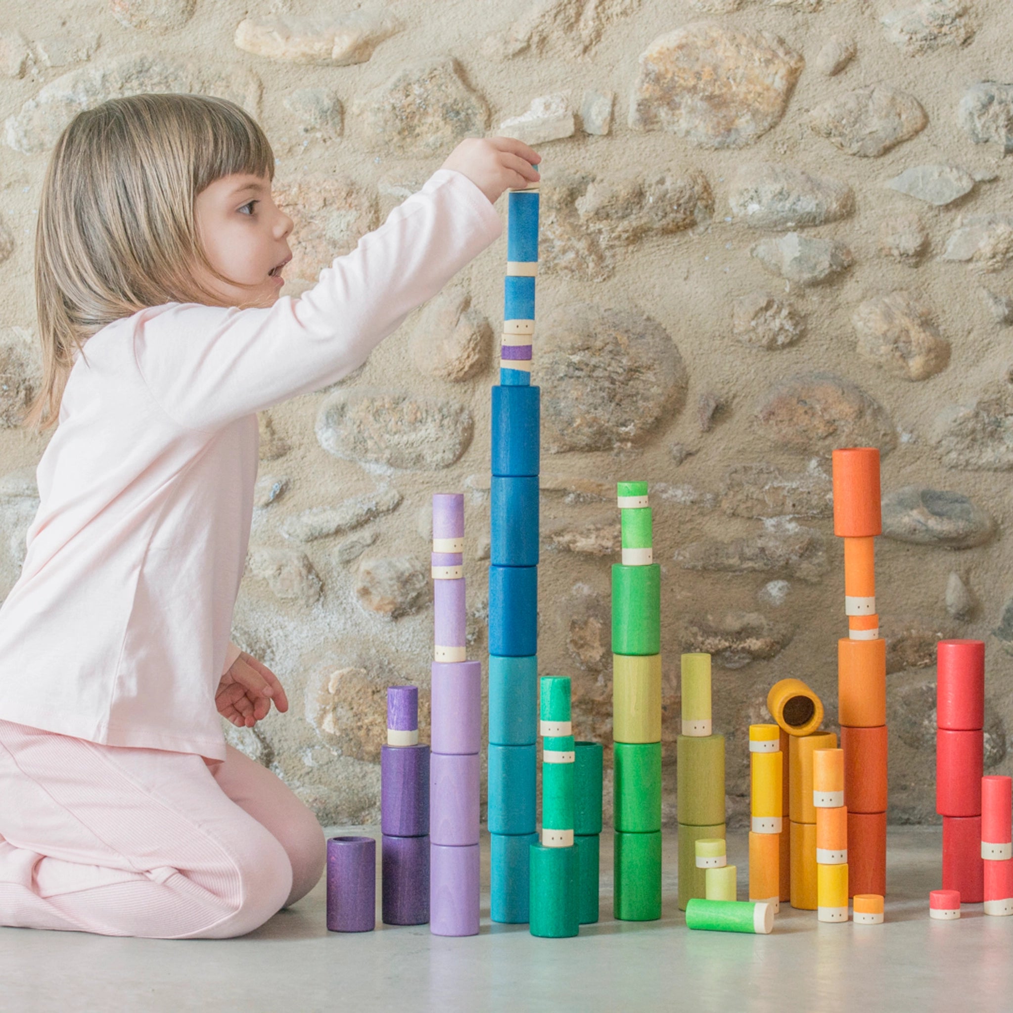 Ein Mädchen spielt mit bunten Holzbausteinen des Sets Freies Spiel: Lola, bestehend aus 72 handgefertigten Teilen aus nachhaltig geforstetem Holz, ideal für kreatives Spielen.