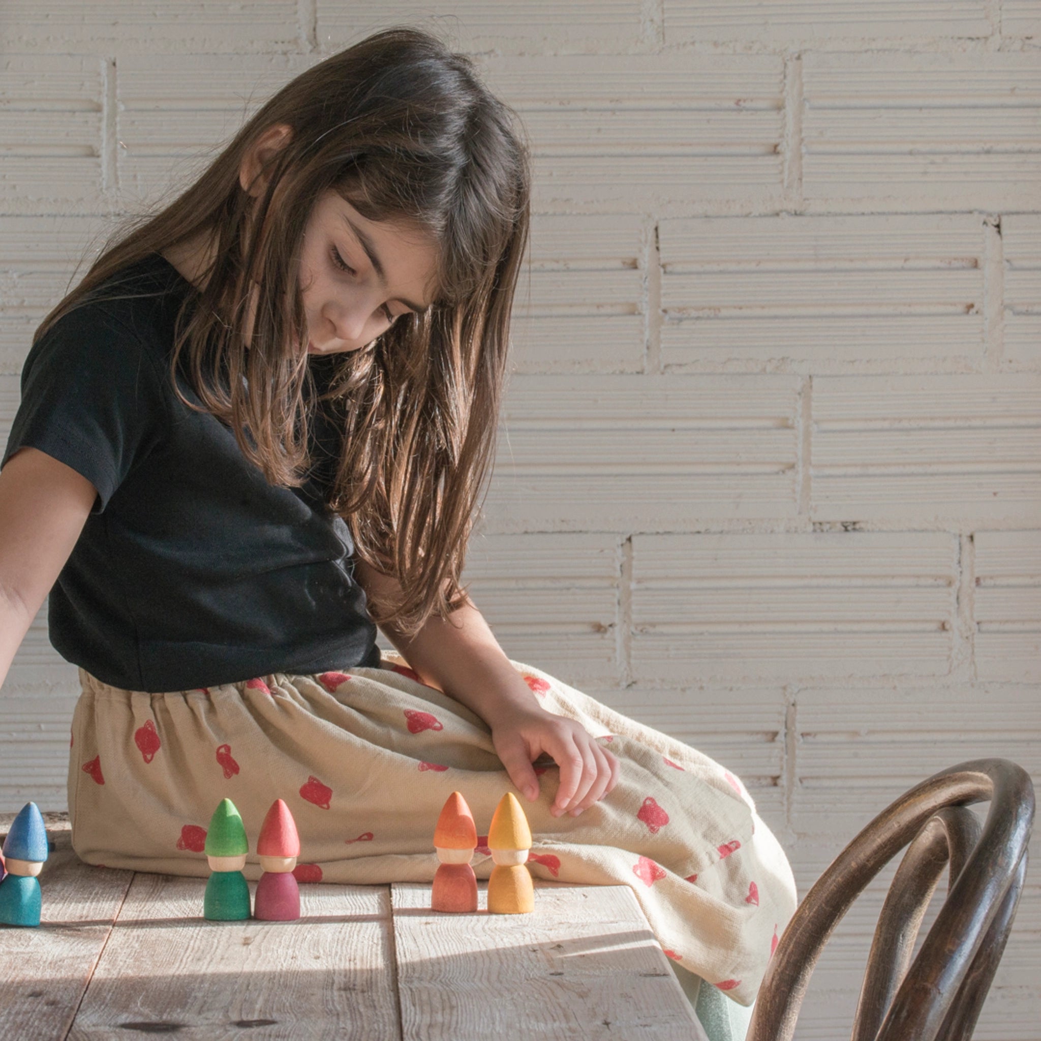 Mädchen spielt am Tisch mit Freies Spiel: Rainbow Tomtens, einem handgefertigten Set aus sechs bunten Zwergfiguren aus nachhaltigem Holz, ideal für kreatives und freies Spiel.