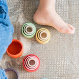 Freies Spiel: Nest Bowls aus handgefertigtem Holz, ideal zum Bauen und Stapeln. Zeigt einen Kinderfuß neben bunten Objekten, einschließlich Holzbechern in verschiedenen Größen und Formen, auf einem Boden liegend.