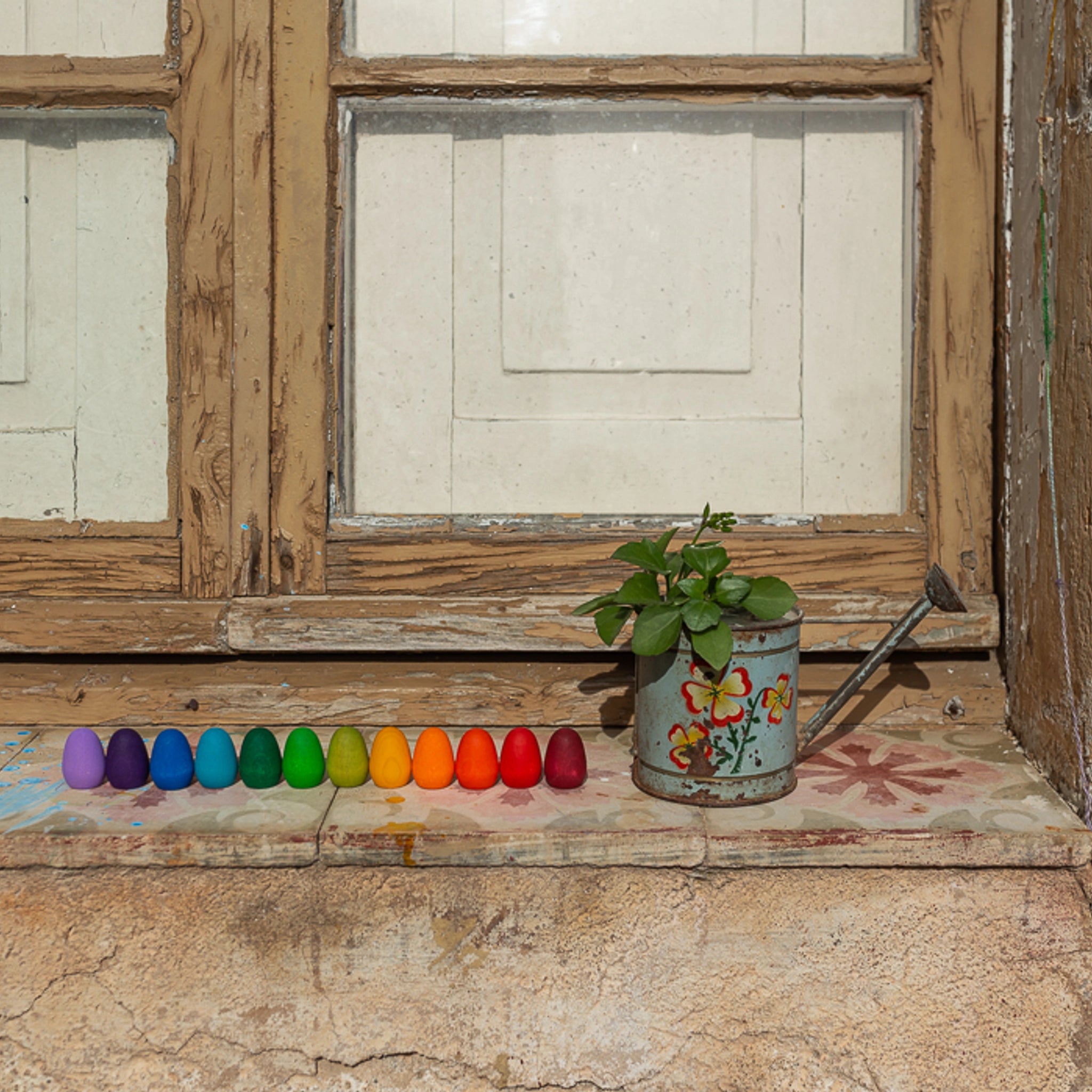 Freies Spiel: Mandala Rainbow Eggs in einem Gießkannen-Arrangement vor einem Fenster. Handgefertigte, nachhaltig produzierte Holzeier für kreative Kinderspiele und bunte Mandalas.