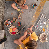 Freies Spiel: Rahmen zeigt zwei Kinder, die auf dem Boden sitzen, umgeben von einem Korb mit Garn und Perlen, ideal für kreatives Spielen und handgefertigt in Spanien aus nachhaltig gewonnenem Holz.