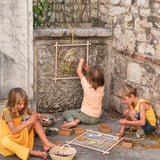 Kinder sitzen draußen an einer Steinmauer, spielen kreativ mit dem handgefertigten Freies Spiel: Frame aus nachhaltigem Holz, ideal zum Weben und Sammeln, inspiriert von KLICKKLACKs ästhetischem und nachhaltigem Designansatz.