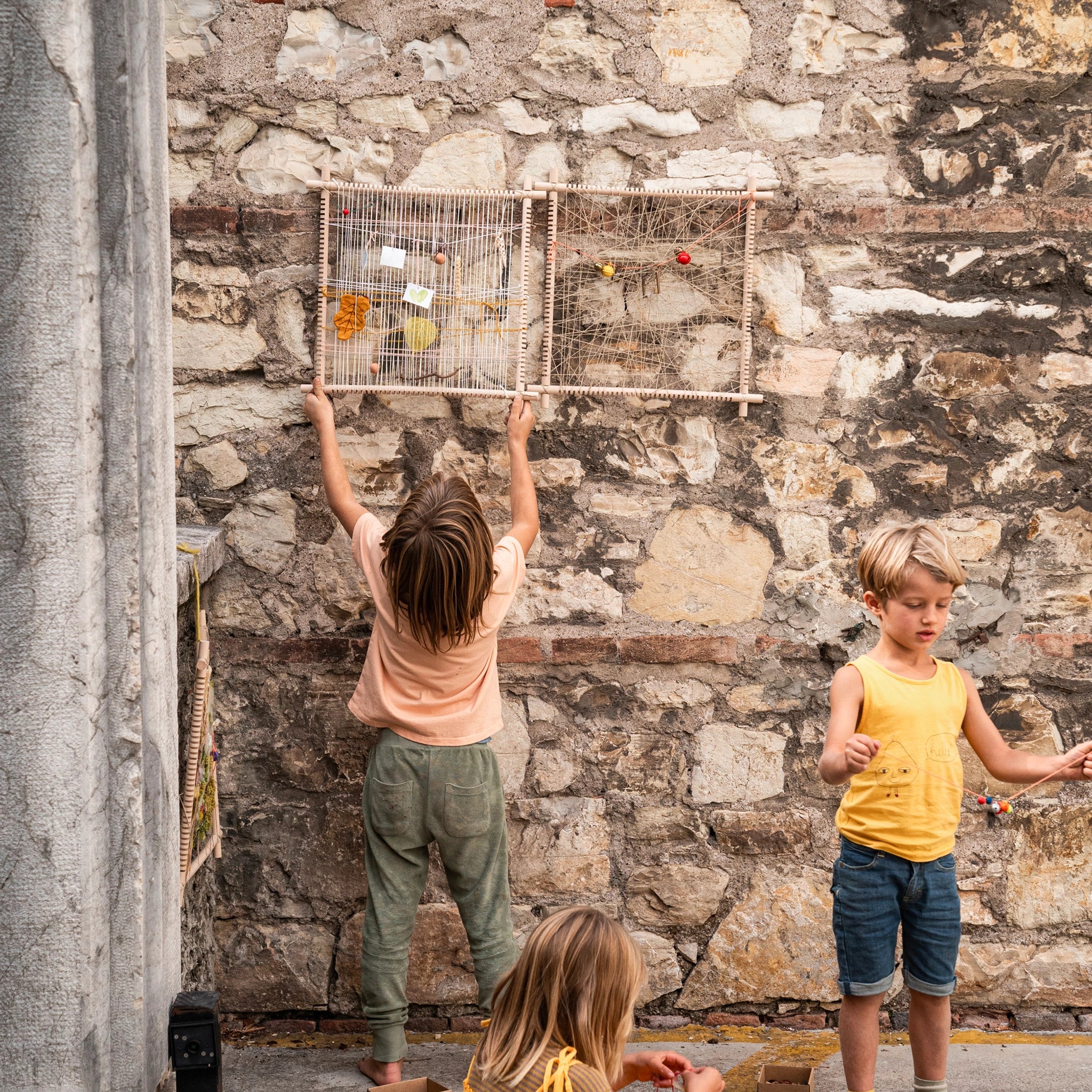 Kinder spielen draußen mit dem Freies Spiel: Frame, einem handgefertigten Holzrahmen aus Spanien, ideal zum Weben und kreativen Spielen, verkörpert nachhaltige Produktion und zeitgemäßes Design von KLICKKLACK.