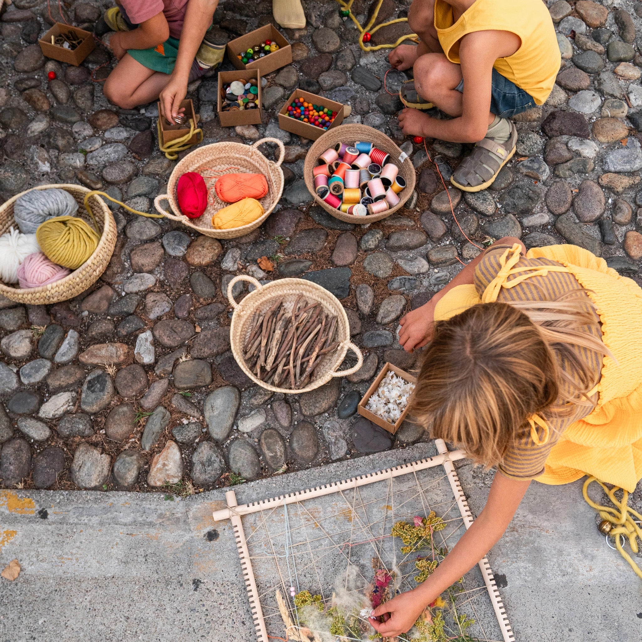 Freies Spiel: Mehrere Kinder sitzen auf einem Steinboden mit Körben voller Garn und Fädenspulen, die den handgefertigten Holzrahmen zum Weben und kreativen Spielen präsentieren. Ideal für nachhaltiges, freies Kinderspiel.