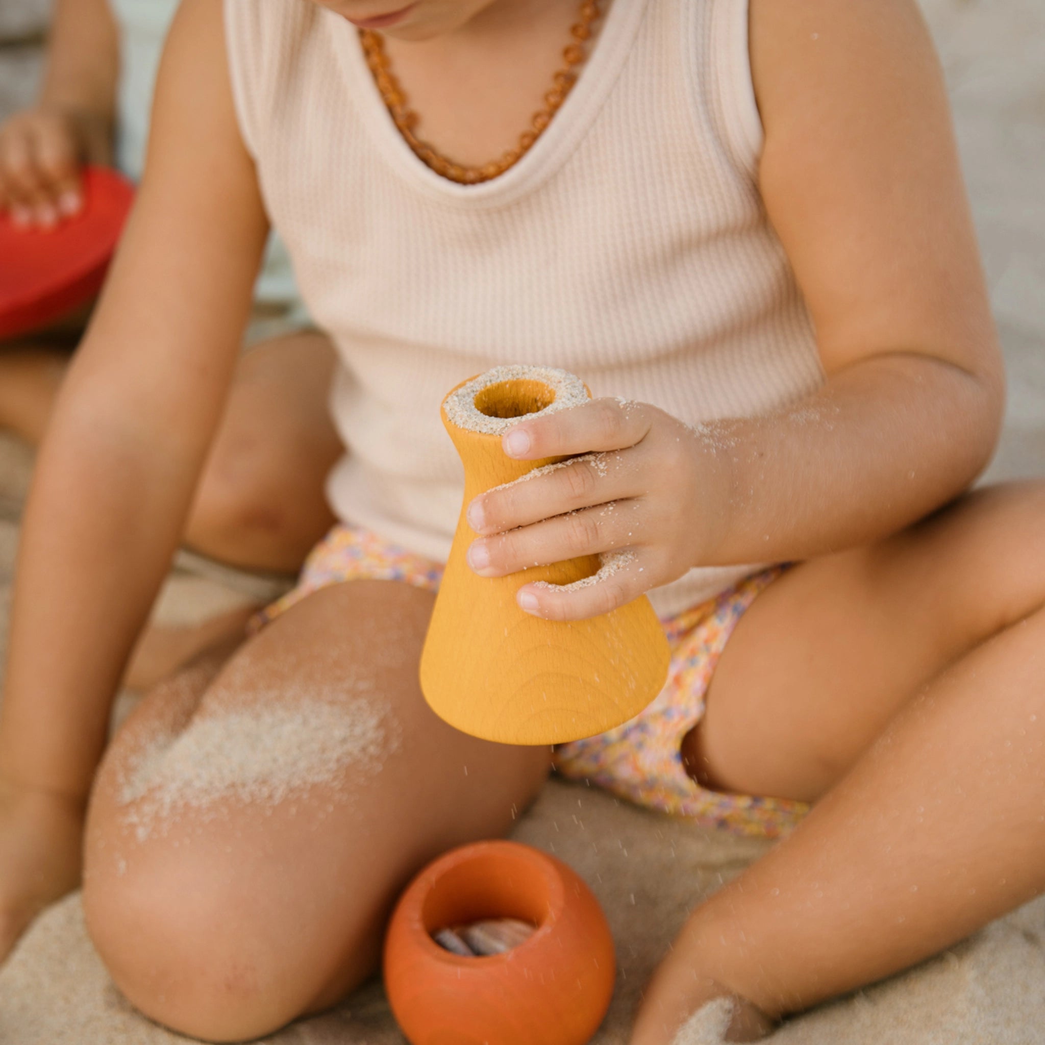 Freies Spiel: Pots aus nachhaltig geforstetem Holz, handgefertigt in Spanien, ideal für Indoor- und Outdoor-Spiele. Ein Kind spielt mit einem der bunten Töpfchen im Sand.