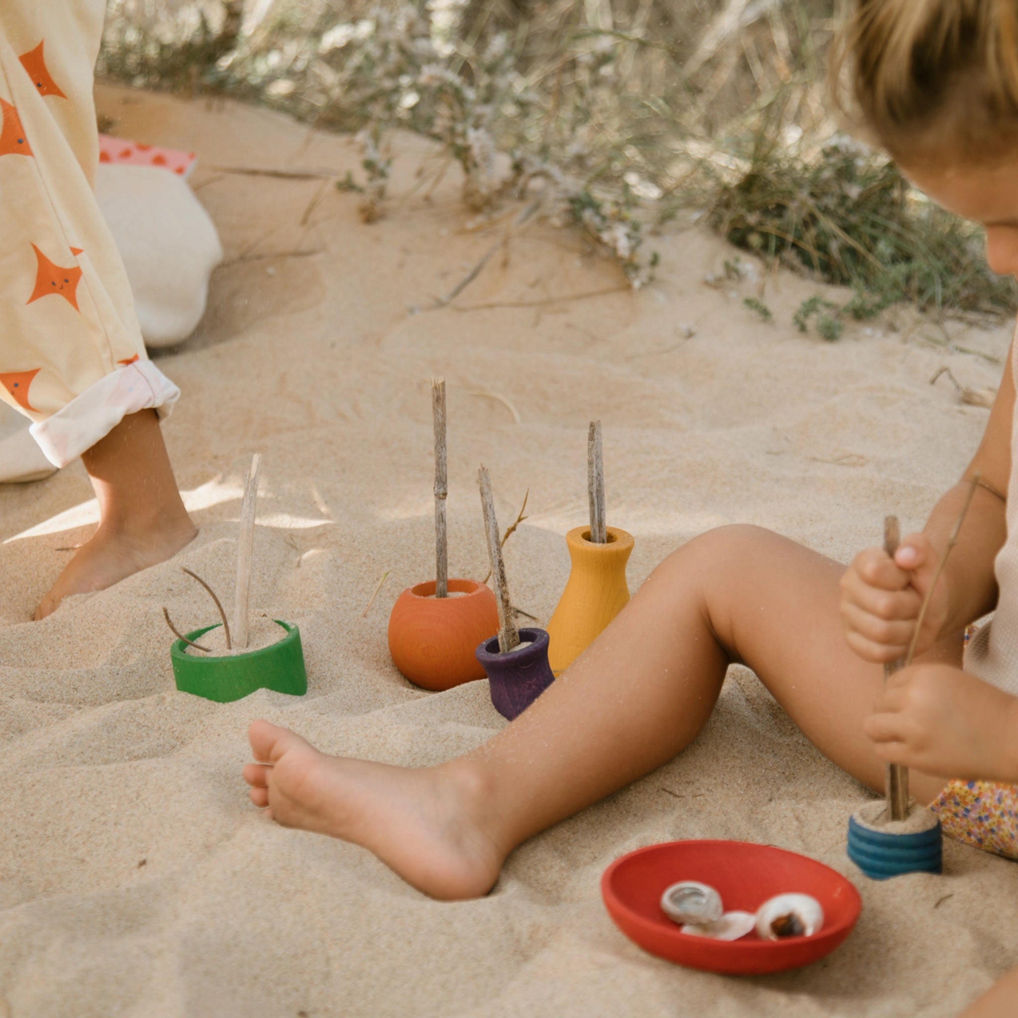 Freies Spiel: Pots aus nachhaltig geforstetem Holz, handgefertigt in Spanien. Ein Kind spielt mit den bunten Holztöpfchen im Sand, ideal für kreatives Indoor- und Outdoor-Spiel.