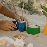 Freies Spiel: Pots aus nachhaltig geforstetem Holz, handgefertigt in Spanien, ideal für drinnen und draußen. Im Bild sind Kinderhände zu sehen, die mit den bunten Holztöpfchen spielen.