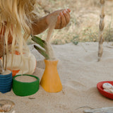 Freies Spiel: Pots - Ein Kind spielt im Sand mit einem grünen Behälter, handgefertigte bunte Töpfchen aus Holz, ideal für kreatives Spielen drinnen und draußen, gefertigt aus nachhaltig geforstetem Holz.