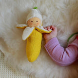 Greifling: Banane, ein kleiner Plüsch-Bananenbaby, liegt auf weicher Oberfläche. Ein Baby hält sanft einen Bananen-Arm. Perfekt für nachhaltige, moderne Ästhetik, ideal für KLICKKLACK's umweltbewusste Kundschaft.