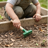 Ein Kind kniet am Holzbeet und benutzt das Kindergartenset, das einen kleinen Handrechen mit grünem Metallkopf umfasst, um den Boden zu bearbeiten.