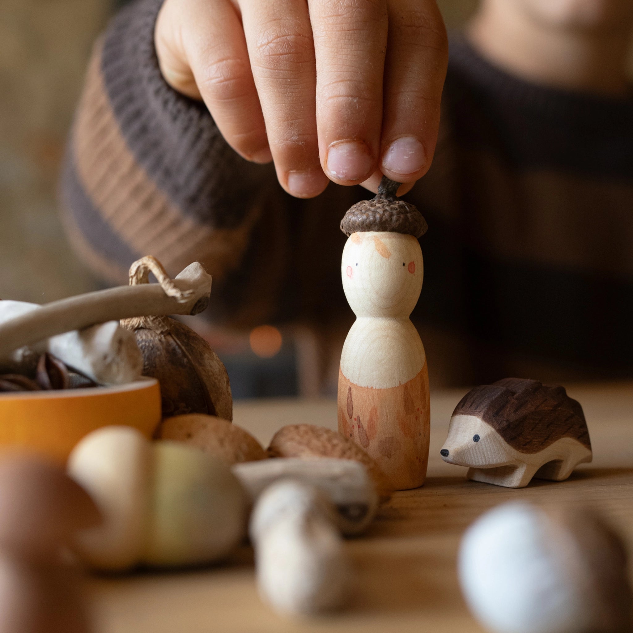 Freies Spiel: Hello autumn! – Ein Kind greift nach einer handgefertigten Holzfigur, dem Eichelmännchen, umgeben von herbstlichen Spielstücken auf einem Tisch. Nachhaltig produziert von Grapat & Ostheimer.