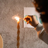 Eine Hand entzündet mit einem Streichholz die Spitze einer hohen, gedrehten Bienenwachskerze. Im Hintergrund: Freies Spiel: Hello winter! Set mit 5 handbemalten Holzelementen, inspiriert von nachhaltiger Produktion.