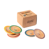 Freies Spiel: Dishes von Grapat, ein Set aus kleinen, runden Holzplatten mit farbigen Rändern und einem zarten Blattmotiv in der Mitte, präsentiert in einer schlichten Pappschachtel.