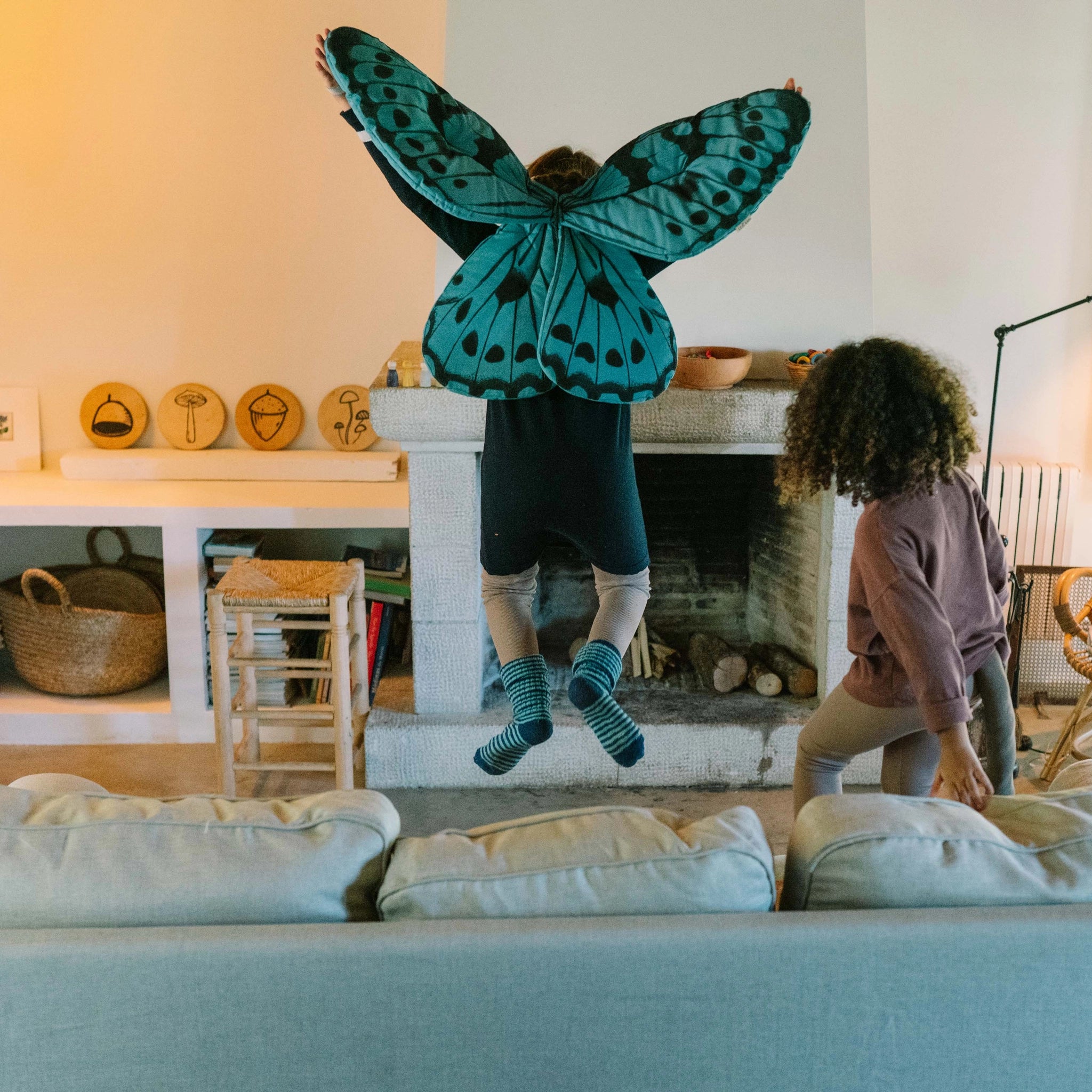Zwei Kinder spielen vor einem Kamin. Eines springt mit Freies Spiel: Warm Mandala Mix Flügeln. Im Vordergrund ein Sofa, im Hintergrund ein Korb und Bücherregal.