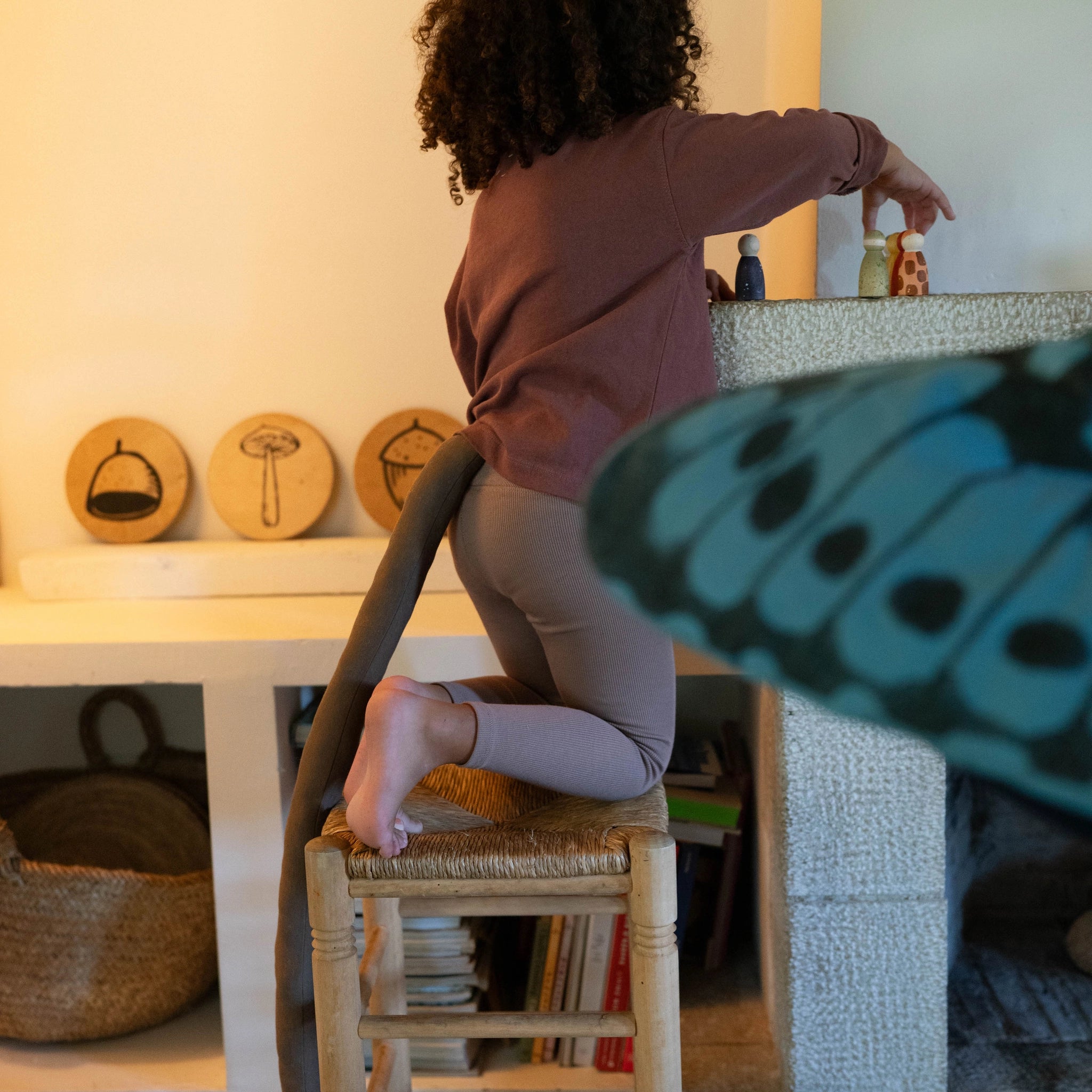 Ein Kind arrangiert die handbemalten Holzfiguren Freies Spiel: Animal Crew auf einem Kaminsims. Die Szene zeigt eine gemütliche, heimelige Umgebung mit gewebten Sitzhockern und dekorativen Holzelementen.