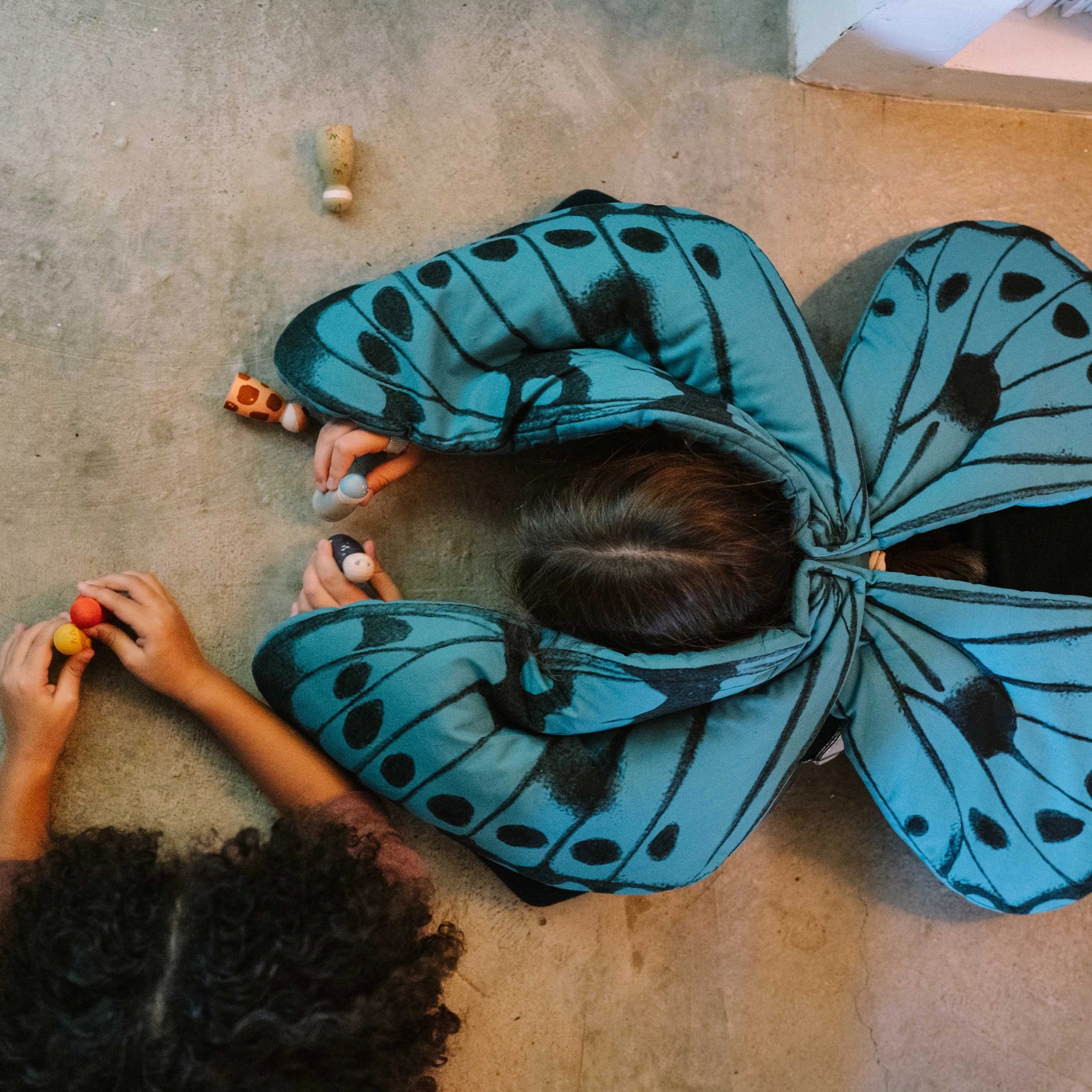 Zwei Kinder spielen auf dem Boden mit bemalten Holzfiguren aus dem Set Freies Spiel: Warm Mandala Mix, die Tier- oder Menschengestalten darstellen. Ein Kind trägt große Schmetterlingsflügel.