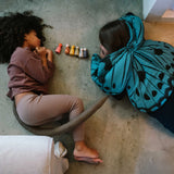 Zwei Kinder spielen auf dem Boden mit Freies Spiel: Warm Mandala Mix, einer Reihe von sechs handbemalten Holzfiguren. Ein Kind trägt einen Schmetterlingsumhang, das andere hat einen weichen Schwanz.