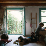 Drei Kinder spielen mit Freies Spiel: Animal Crew auf einem warmen Holzboden in einem sonnendurchfluteten, rustikalen Raum. Zwei Kinder arrangieren die hölzernen Spielfiguren, während das dritte ein leuchtendes Objekt hält.