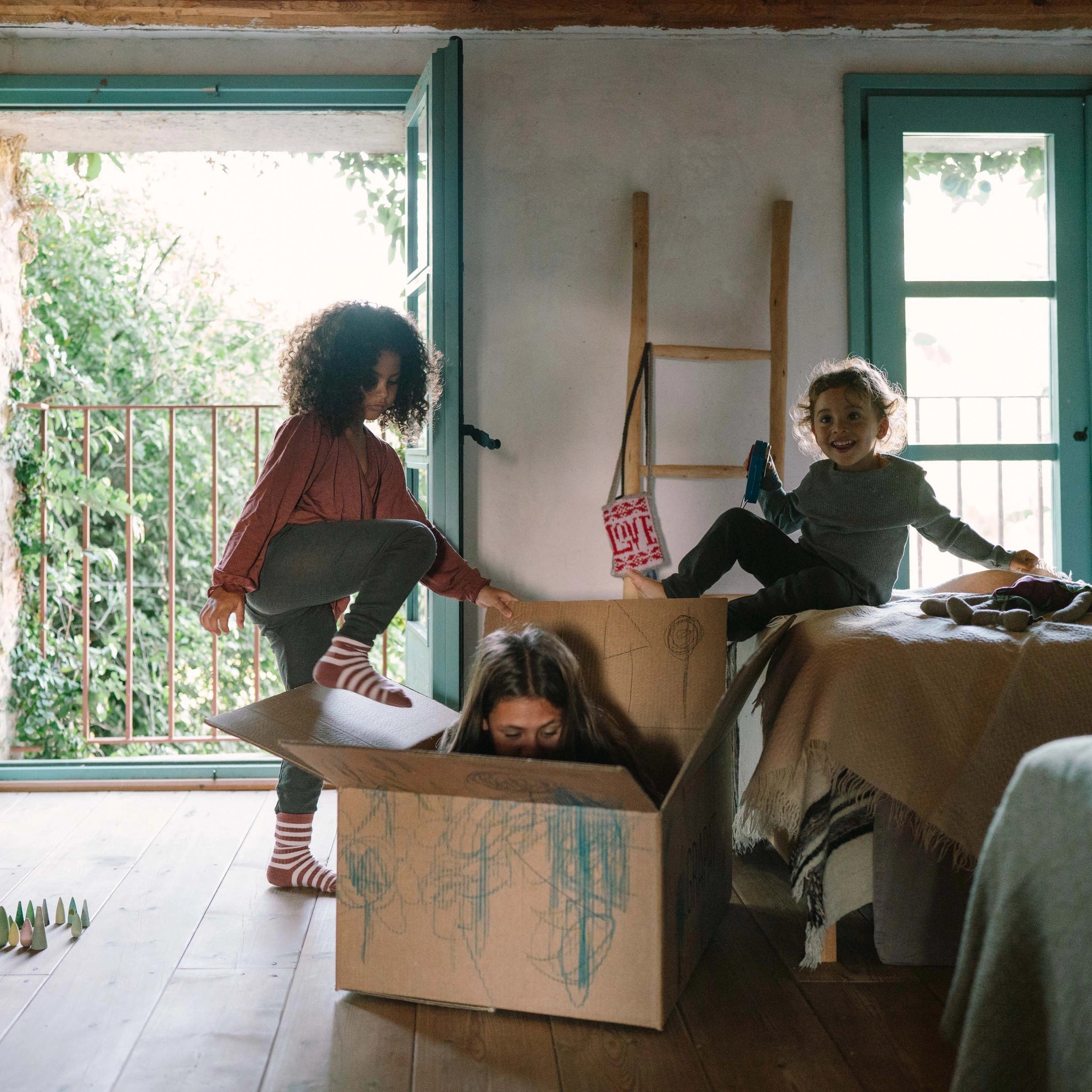 Kinder spielen mit Freies Spiel: Animal Crew in einem sonnigen Raum. Ein Kind steht auf einem großen Karton, ein anderes kniet darin, während ein drittes Kind auf dem Bett mit einem Gadget lächelt.