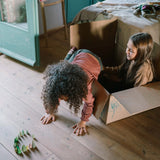 Zwei Kinder spielen mit dem Freies Spiel: Animal Crew. Eines sitzt in einem Karton, das andere krabbelt heraus. Vor ihnen stehen grüne Holzfiguren, die kreative Fantasie fördern.