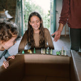 Drei Kinder spielen freudig mit dem Freies Spiel: Animal Crew Set, bestehend aus kleinen, holzgeschnitzten Bäumen und einer Schnecke, arrangiert auf einem Karton zu einem improvisierten Waldspiel.