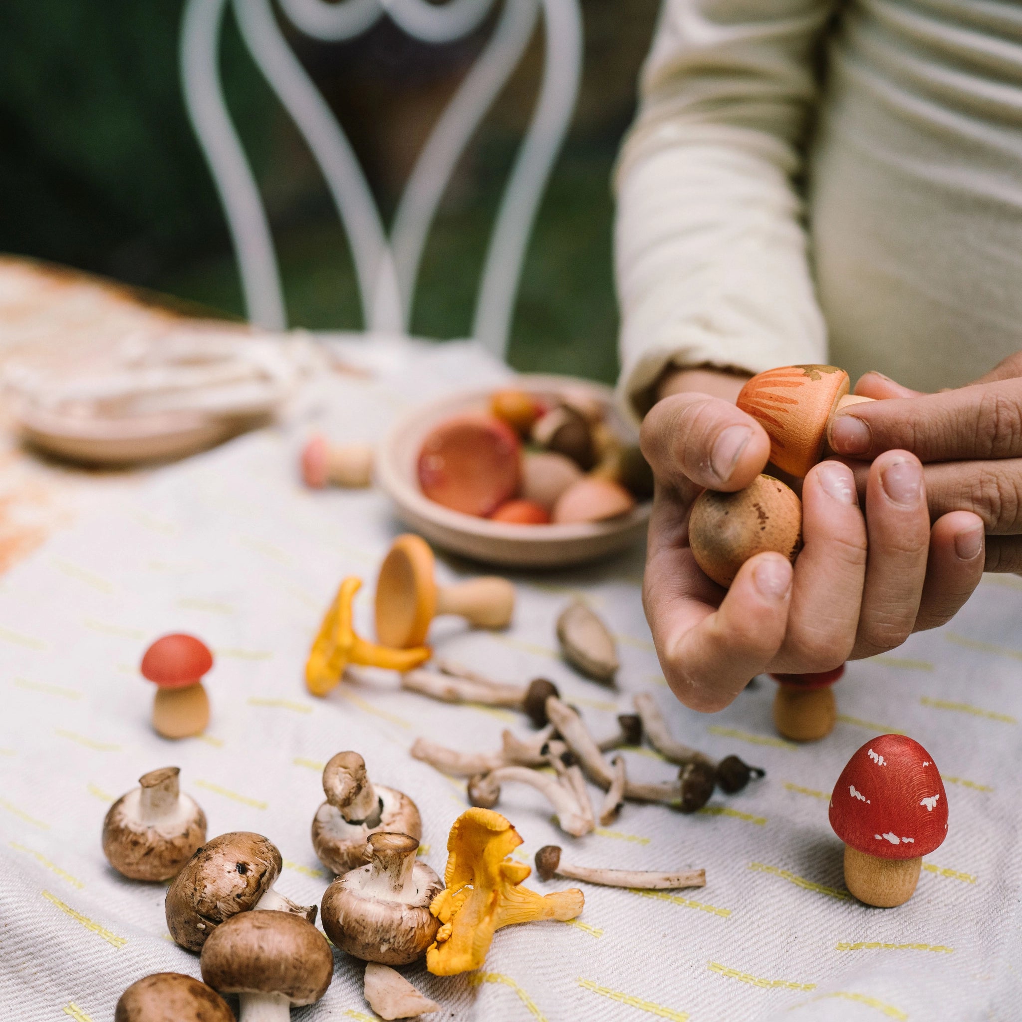Freies Spiel: Forest Tree – Auf einem mit sanften Streifen gemusterten Tuch liegen echte Pilze und handbemalte Holzspielzeugpilze, die von einer Person gehalten werden. Ein hölzernes Schälchen enthält weitere Spielpilze.