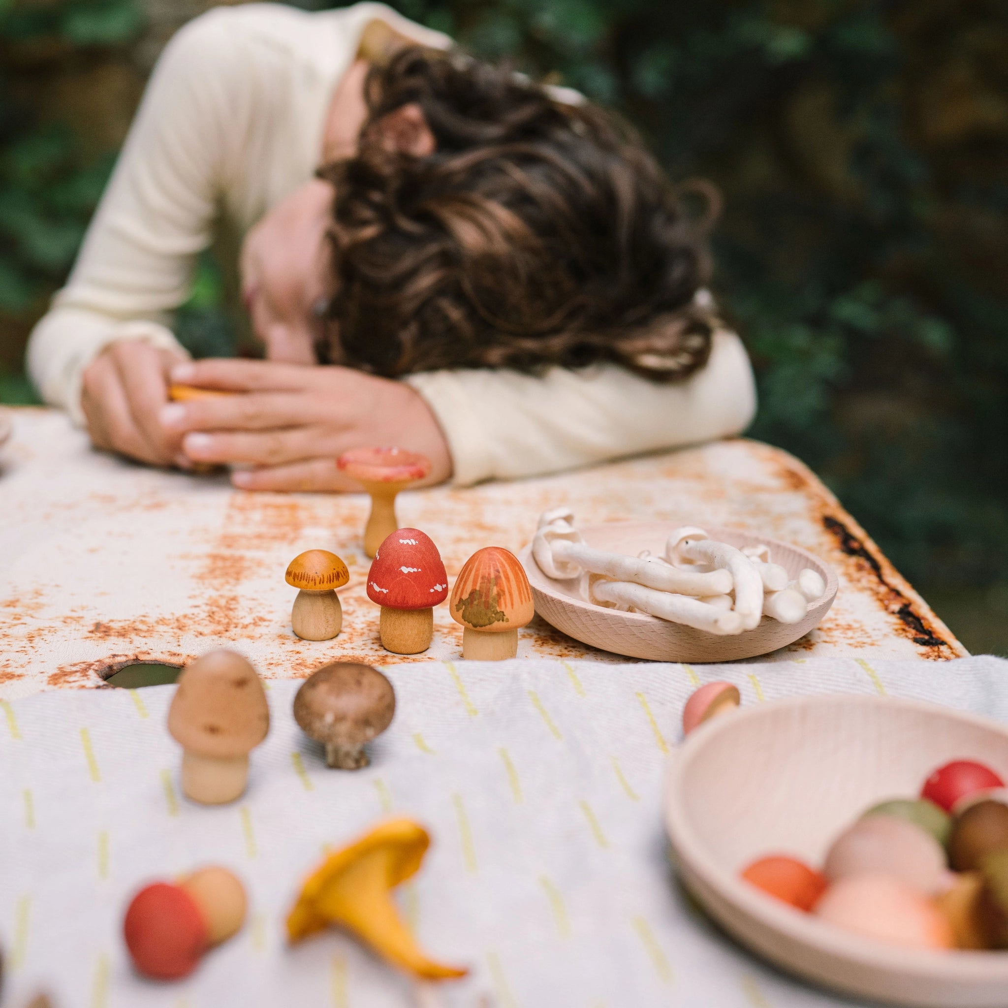 Ein Kind lehnt sich über einen verwitterten Tisch und hält einen kleinen Gegenstand. Umgeben von handbemalten und realistischen Pilzen liegt das Freies Spiel: Mushroom Grove Set mit Holzschalen und Stoff.