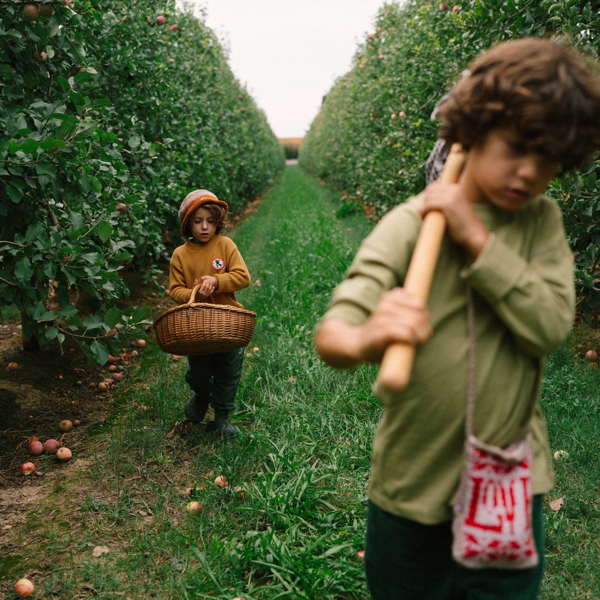 Zwei Kinder im Apfelgarten: Das jüngere Kind trägt für das Freies Spiel: Fruits einen Korb, das ältere eine Stange zum Pflücken. Ideal für nachhaltiges, kreatives Spielen im Freien.