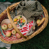 Ein geflochtener Korb enthält Freies Spiel: Fruits, einschließlich handbemalter Holzfrüchte und -spielstücke in einer Schale, ergänzt durch handgestrickte Hülle und Tuch, ideal für kreatives, nachhaltiges Spielen.
