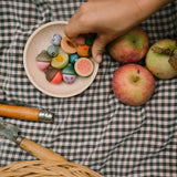 Freies Spiel: Fruits – Ein flacher Holzteller voller kleiner, leuchtend bemalter Holzfrüchte auf einem Karo-Tuch, mit einer Hand, die ein Stück greift. Drei Äpfel, ein Klappmesser und ein Gartenwerkzeug liegen daneben.