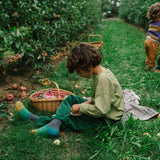 Zwei Kinder im Apfelgarten, eines sitzend mit Freies Spiel: Fruits, in grünen Cordhosen und gestrickten Socken, neben einem Korb mit Äpfeln. Ein weiteres Kind pflückt Äpfel im Hintergrund.