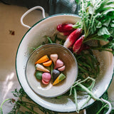 Ein weißes Emaille-Sieb von Freies Spiel: Veggies, gefüllt mit frischem Gartengemüse und einer Schale kleiner, bunter Holzspielzeug-Gemüse, passend zur ästhetischen und nachhaltigen Philosophie von KLICKKLACK.