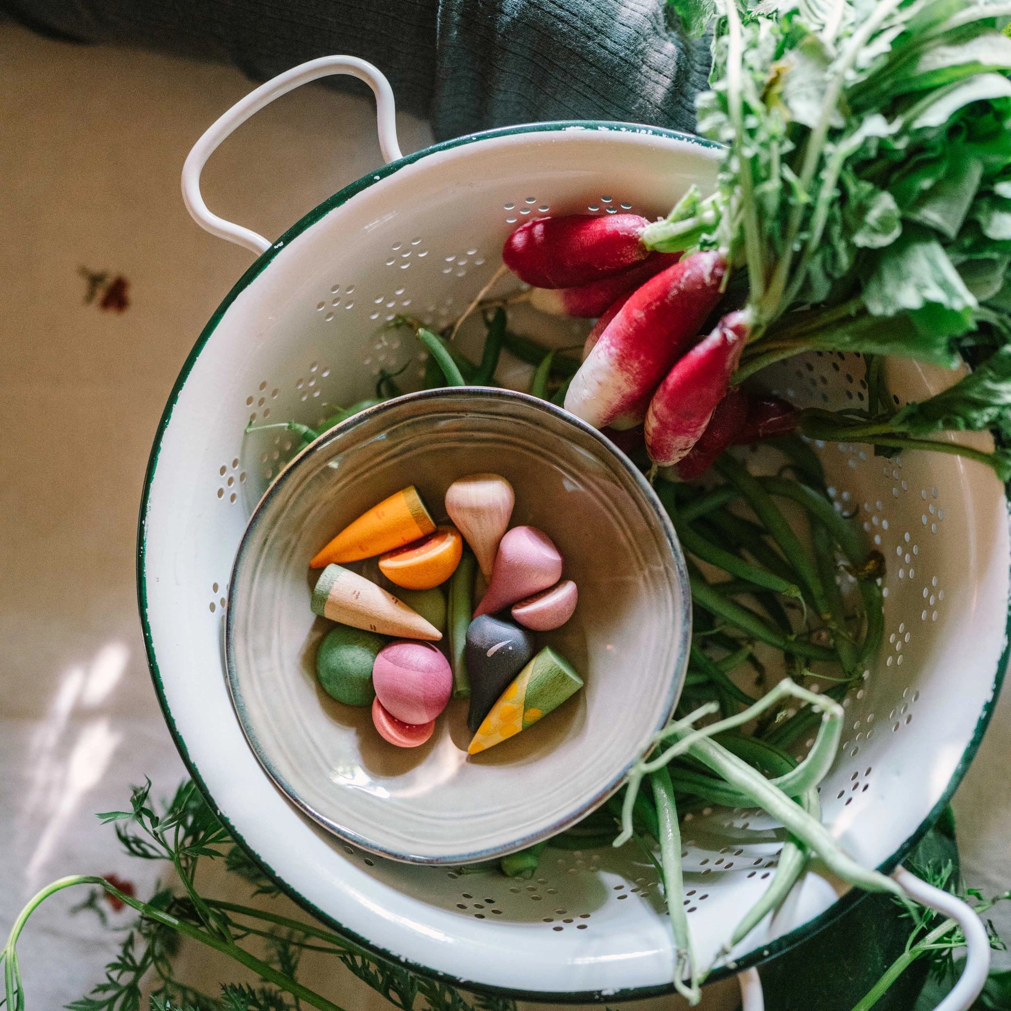 Ein weißes Emaille-Sieb von Freies Spiel: Veggies, gefüllt mit frischem Gartengemüse und einer Schale kleiner, bunter Holzspielzeug-Gemüse, passend zur ästhetischen und nachhaltigen Philosophie von KLICKKLACK.