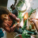 Freies Spiel: Veggies zeigt eine gemütliche Küchenszene, in der ein Kind Karotten schält und mit einem Helfer frisches Gemüse auf einem Holzbrett vorbereitet, ideal für nachhaltige Familienküchen.