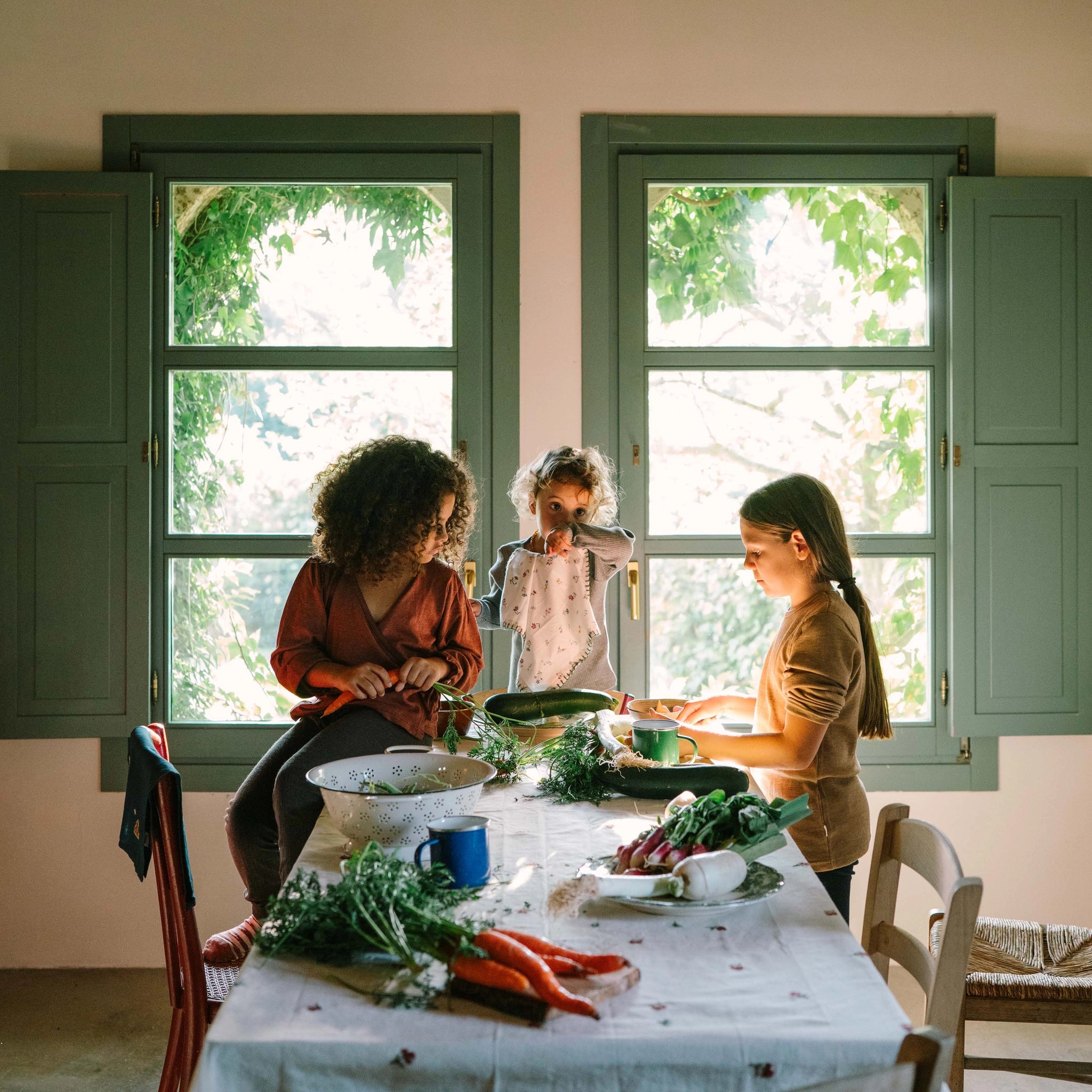 Drei Kinder um einen rustikalen Holztisch im weichen Tageslicht, bearbeiten frisches Gemüse. Titel: Freies Spiel: Veggies. Tisch steht vor großen Fenstern, symbolisiert nachhaltige Freude und regionale Produktion.