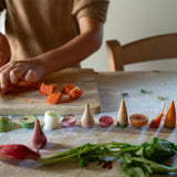Freies Spiel: Veggies – Hände schneiden Karotten auf einem Holzbrett, umgeben von halben, spielzeugartigen Gemüsemodellen wie Radieschen, Lauch und Avocado, die KLICKKLACKs nachhaltige und ästhetische Produktphilosophie verkörpern.