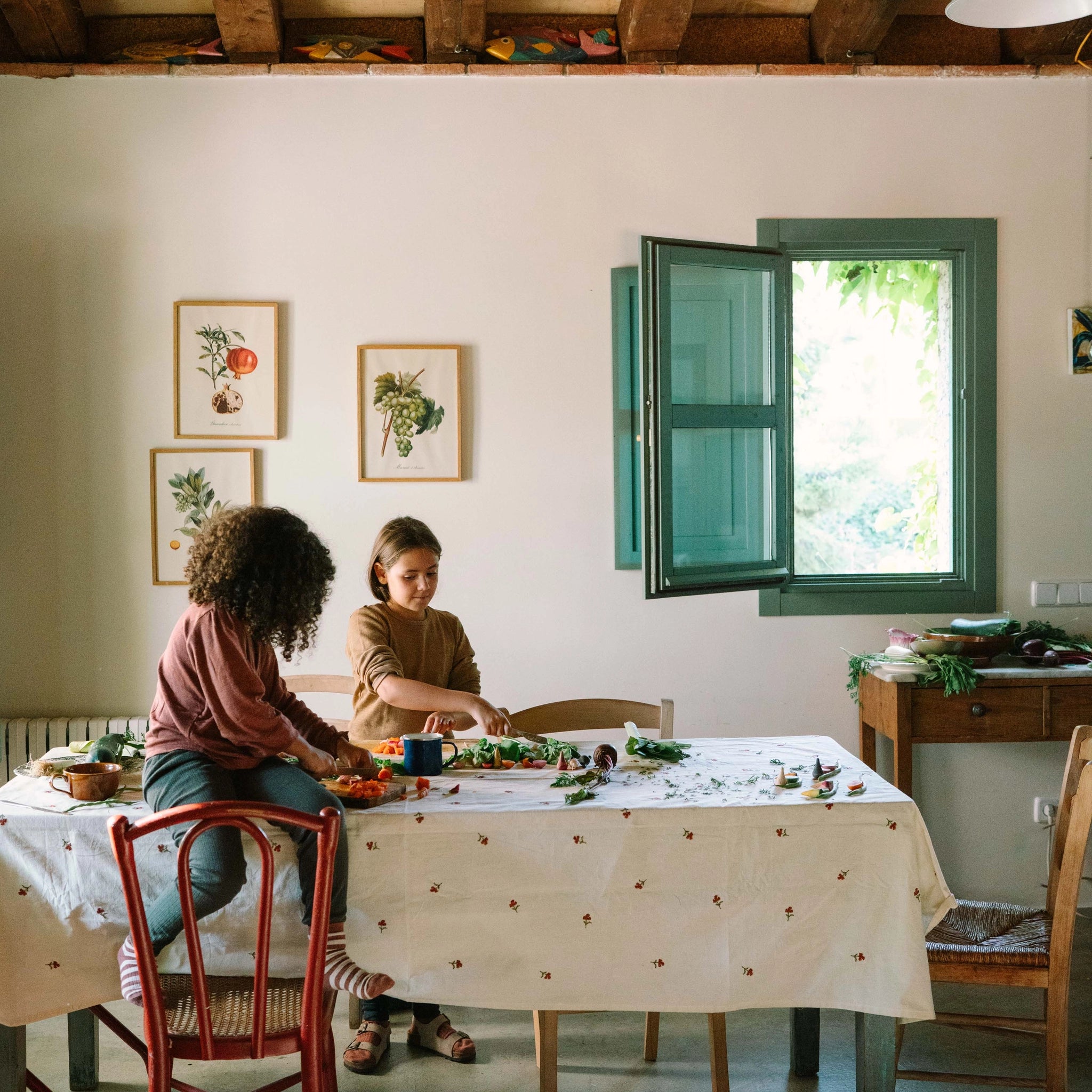 Freies Spiel: Veggies – Zwei Kinder bereiten frisches Gemüse an einem großen Tisch mit Blumenmuster-Tischdecke vor. Der Raum strahlt eine gemütliche Atmosphäre mit botanischen Drucken und natürlichem Licht aus.