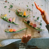 Ein Kind spielt mit Freies Spiel: Veggies. Vier grüne Bohnenboote tragen bunte Holzgemüse wie Karotte, Mais, Gurke, Tomate und Rübe, dekoriert mit grünen Blättern, auf einem neutralen Tuch.