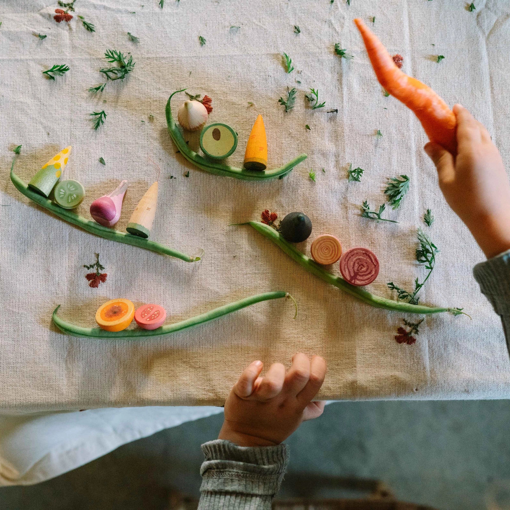 Ein Kind spielt mit Freies Spiel: Veggies. Vier grüne Bohnenboote tragen bunte Holzgemüse wie Karotte, Mais, Gurke, Tomate und Rübe, dekoriert mit grünen Blättern, auf einem neutralen Tuch.