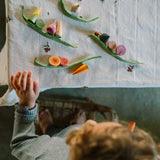 Ein Kind greift nach dem Freies Spiel: Veggies, einem Set aus konischen und scheibenförmigen Holzspielzeug-Gemüsen und -Fruchtscheiben, arrangiert auf einem Tisch mit bestickter Tischdecke, während es auf einem Hocker steht.