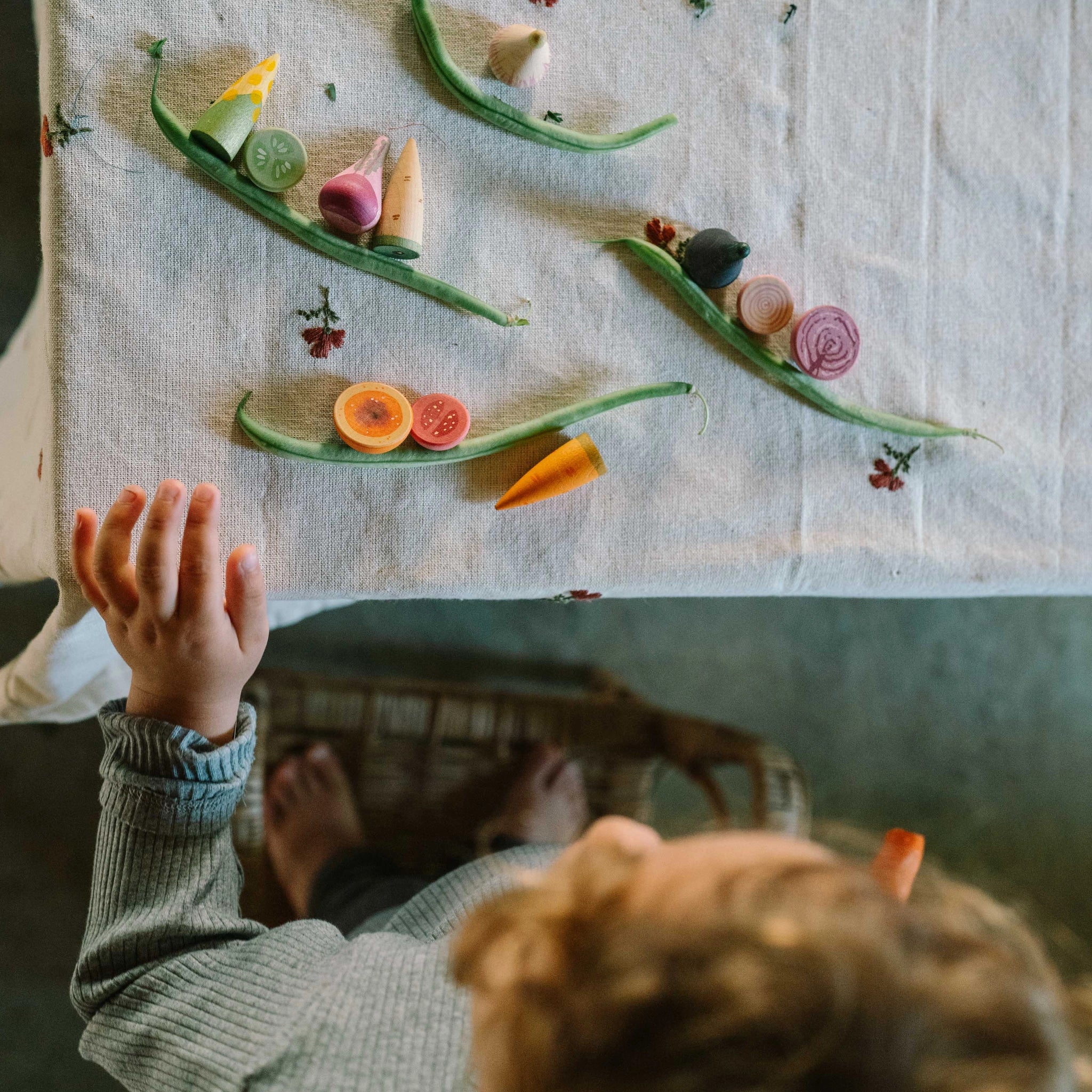 Ein Kind greift nach dem Freies Spiel: Veggies, einem Set aus konischen und scheibenförmigen Holzspielzeug-Gemüsen und -Fruchtscheiben, arrangiert auf einem Tisch mit bestickter Tischdecke, während es auf einem Hocker steht.