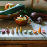 Freies Spiel: Veggies - Handbemalte Holzspielstücke in Form von Gemüse und Früchten auf einem Marmorsockel, umgeben von echtem Gemüse auf der Küchenarbeitsplatte, passend zum nachhaltigen und ästhetischen Ansatz von KLICKKLACK.