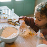 Ein Kind spielt mit Freies Spiel: Bakes, seine Hände in Mehl getaucht, während es Teig mit Ausstechern formt. Ein Tisch voller Mehl und geformter Teigstücke kennzeichnet das Backabenteuer.
